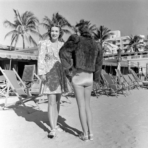 Two women stand on a sandy beach with empty lounge chairs and palm trees in the background. One wears a dress and smiles at the camera, while the other has a fur jacket and swimsuit, facing away. Buildings are visible behind them.