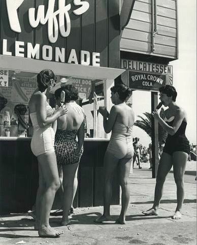 Four women in vintage swimsuits stand at a food stand labeled "Puff's Lemonade" and "Delicatessen," enjoying drinks on a sunny beach day. A Royal Crown Cola sign and palm tree are visible in the background.