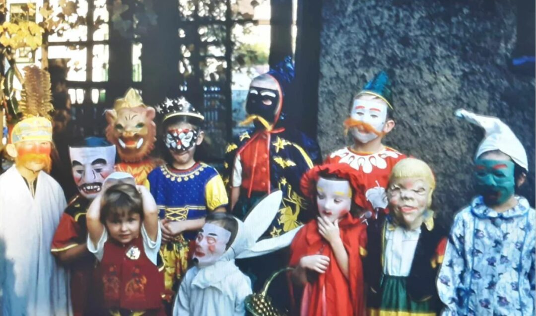 A group of children dressed in colorful costumes and masks, some with painted faces, pose outdoors near a large tree. The costumes include animals, clowns, fairytale, and fantasy characters.