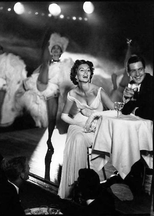 A glamorous woman in a long gown and gloves sits at a small table with a man, both smiling, while cancan dancers perform energetically on stage behind them in a lively, vintage nightclub scene.