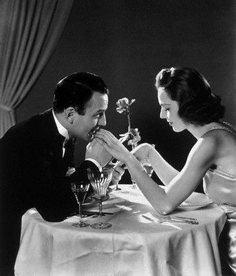 A man in a suit kisses a woman's hand across a candlelit dinner table, with a single rose in a vase between them. Both appear elegant and focused on each other in a romantic, classic black-and-white setting.