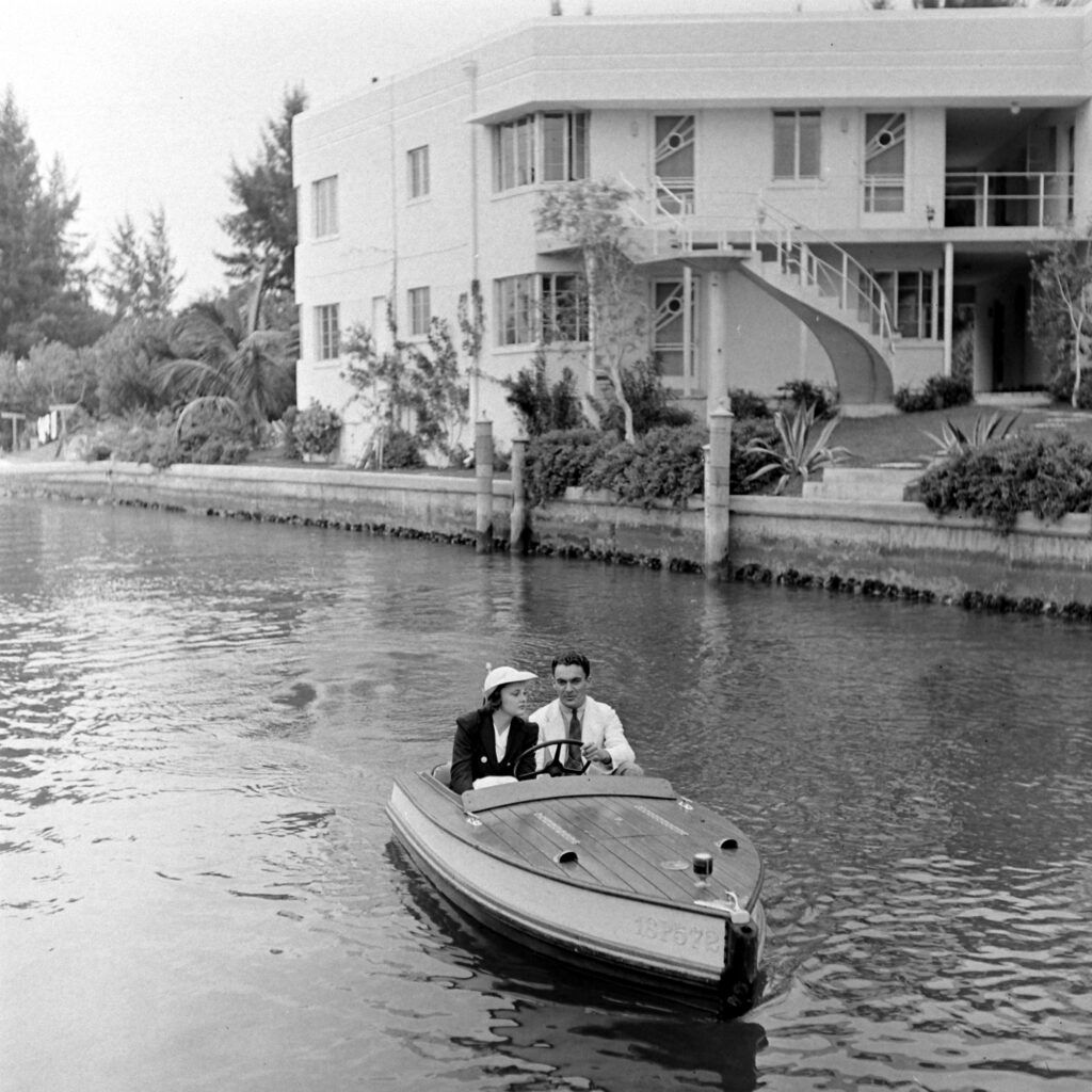 A man and woman in formal clothing ride a small motorboat along a canal, with a modern two-story white building and lush greenery in the background.