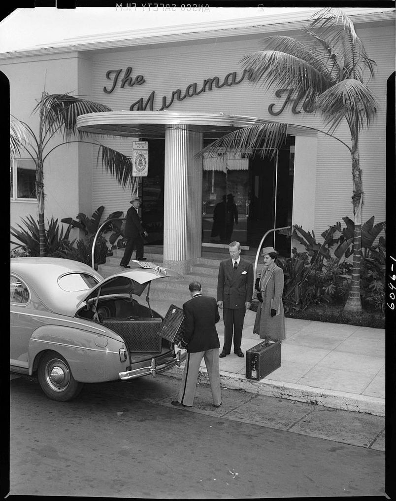 A man loads luggage into the trunk of a vintage car parked in front of The Miramar Hotel, while two guests in formal attire stand on the sidewalk under palm trees, circa 1940s.