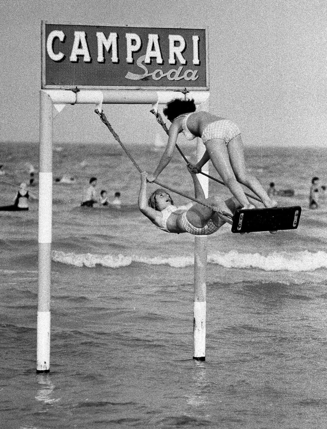 Two women in swimsuits play on a swing set partially submerged in the sea under a sign reading "CAMPARI Soda," with people swimming and waves in the background. The photo is in black and white.