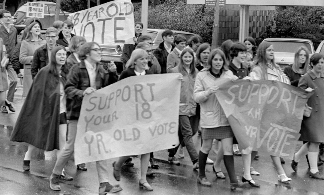 Black and white photo of a group of young people marching in the street, holding signs that say “Support your 18 yr. old vote” and “Support the 18-year-old vote.” Cars are parked along the street in the background.