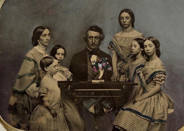 A vintage sepia photograph of a man seated at a table, surrounded by five women and girls in Victorian-era dresses, all posing solemnly for the camera.