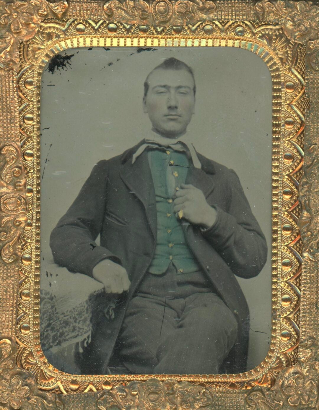 A vintage portrait of a man with short hair, wearing a dark jacket, green vest, and necktie, sitting with one hand on his chest. The photo is framed in an ornate, gold-patterned case.