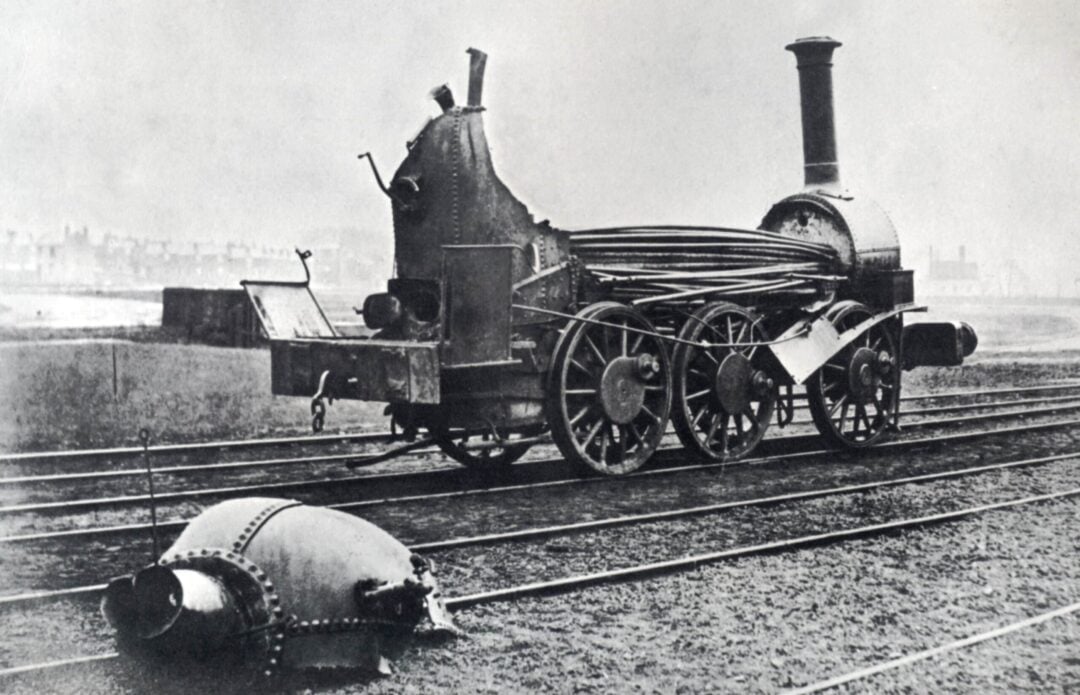 A black-and-white photo of a steam locomotive with a damaged boiler, sitting on railway tracks. A large detached section of the boiler lies on the ground in front of the train. Buildings are visible in the distant background.