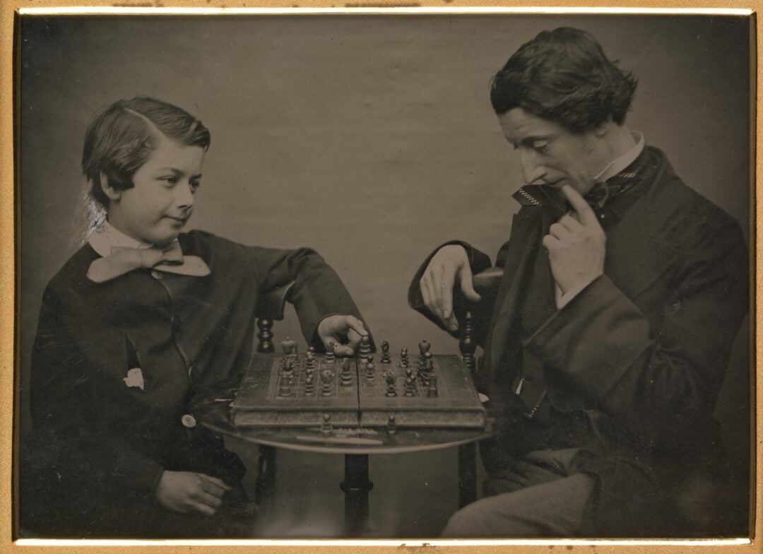 A vintage photograph shows a young boy and an adult man playing chess at a small table. The boy smiles while making a move, and the man contemplates his next move, holding his hand to his face in concentration.