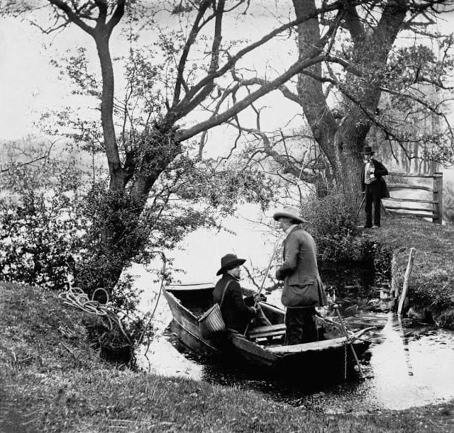 Two people wearing hats sit in a small rowboat at the edge of a wooded riverbank, while a third person stands nearby on the grassy shore, watching them. The scene is in black and white.