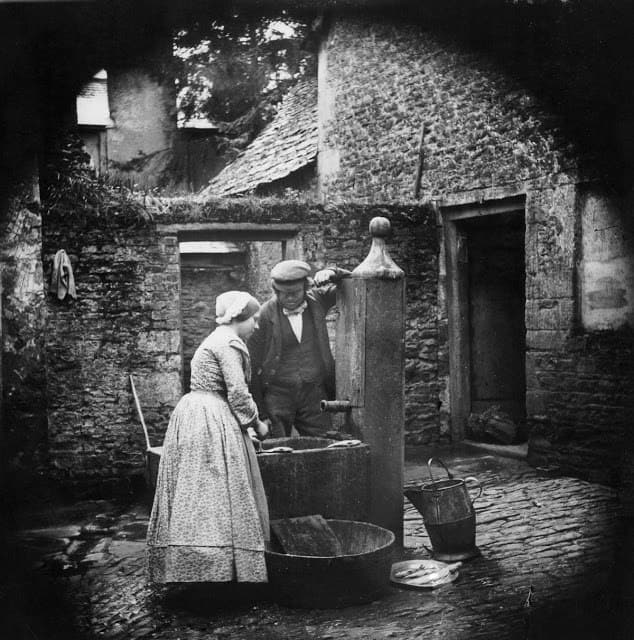 A woman in a long dress and bonnet and a man in a cap draw water from an outdoor stone pump in a cobbled courtyard, surrounded by old stone buildings, buckets, and a wooden tub.