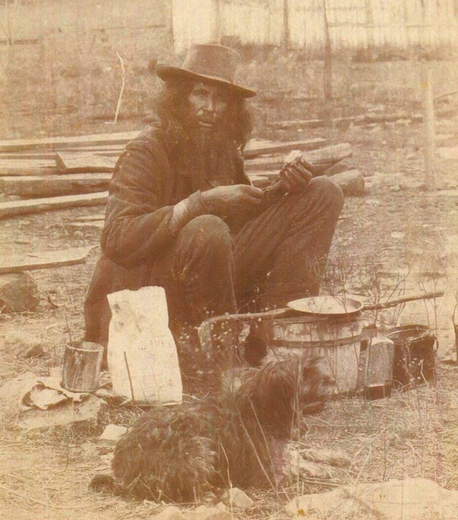 A bearded man in worn clothes and a hat squats outdoors, holding a loaf of bread near cooking pots and supplies; a small shaggy dog lies on the ground in front of him, with a wooden fence and buildings in the background.