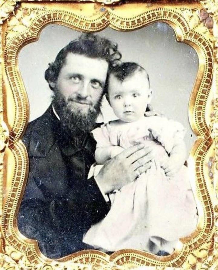 A vintage photograph in an ornate gold frame shows a bearded man in a suit holding a baby in a light dress, both looking at the camera with neutral expressions.