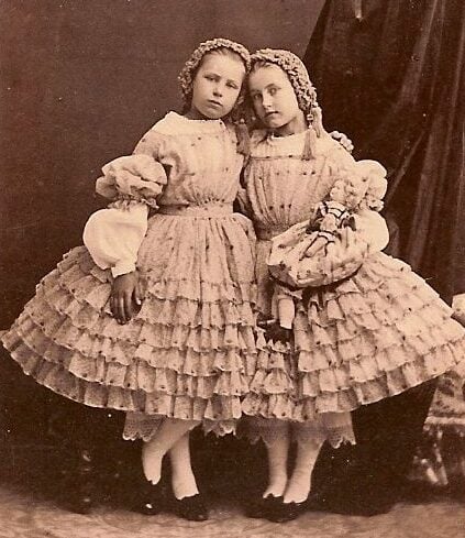 Two young girls in frilly, layered dresses and bonnets stand close together, one resting her head on the other's shoulder. They pose against a dark studio backdrop in a vintage, sepia-toned photograph.