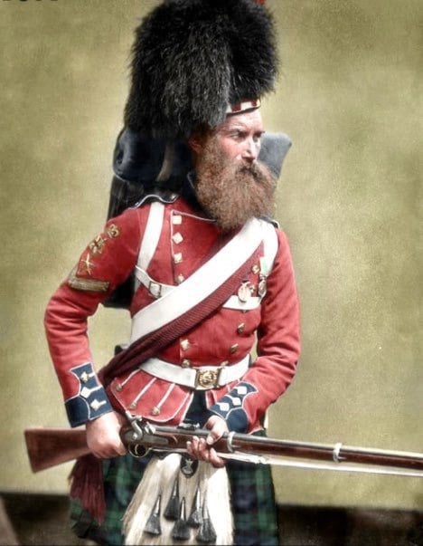 A bearded man in a historic British military uniform with a tall bearskin hat, red jacket, plaid kilt, and white crossbelts holds a musket.