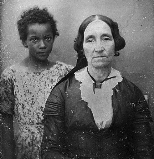 A black-and-white vintage photograph shows a young Black girl standing behind a seated older white woman dressed in dark 19th-century clothing with a lace collar. Both have serious expressions and face the camera.