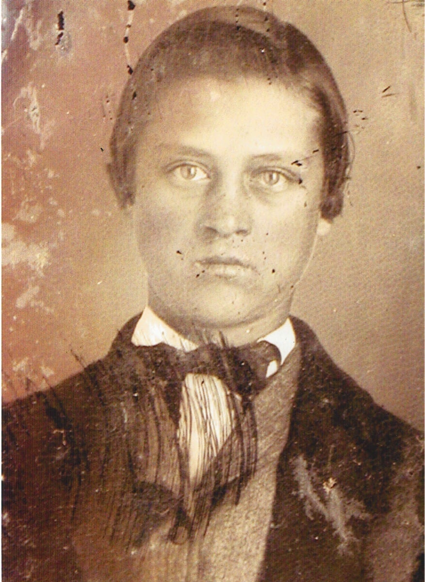 Sepia-toned, old photograph of a young man with short hair, wearing a suit, white shirt, and bow tie. The image is aged and has visible scratches and spots. The young man looks directly at the camera.