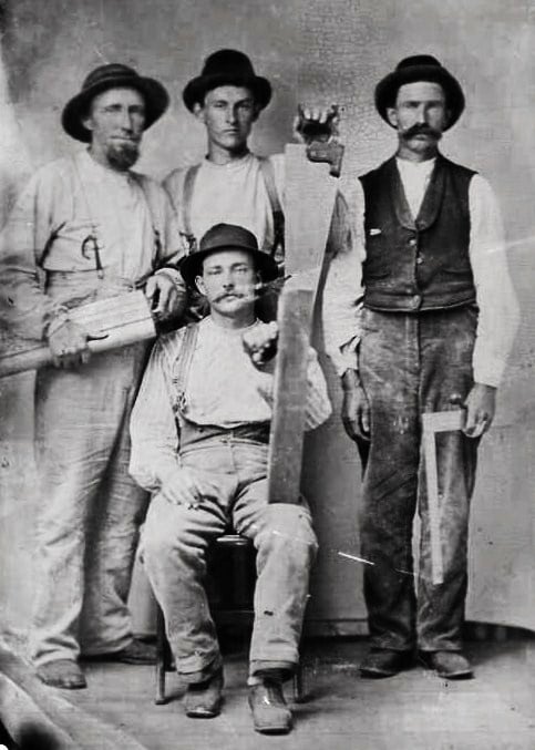 Four men in old-fashioned work clothes and hats pose for a black-and-white photo. Three stand while one sits, and they hold large saws and hand tools, suggesting they are lumberjacks or carpenters.