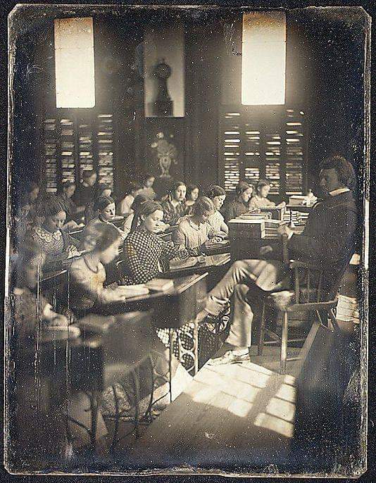 A sepia-toned, vintage photograph shows a classroom with rows of students at desks writing, while a teacher sits at the front. Sunlight streams through tall windows, casting shadows on the wooden floor.