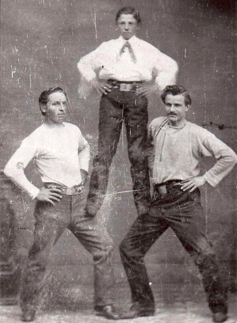 Three men pose in a vintage black-and-white photo; two stand with hands on hips while the third stands balanced atop their thighs, supported by their shoulders. The image appears old and slightly scratched.