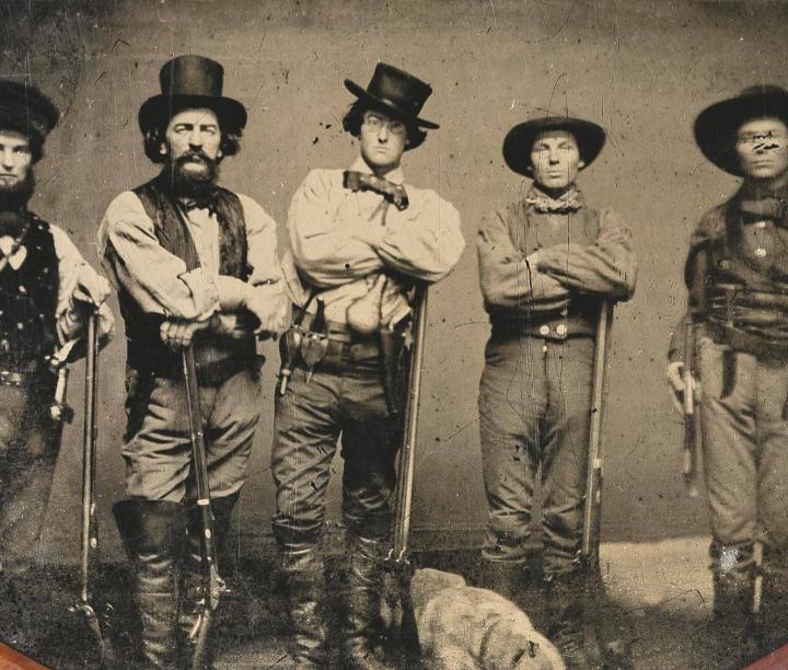 Five men in 19th-century cowboy attire stand side by side, holding long rifles. They wear wide-brimmed hats, boots, and vests, with serious expressions, posing for an old sepia-toned photograph.