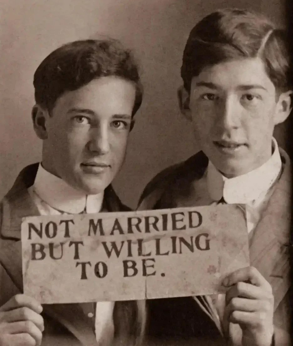 Two young men in early 20th-century attire pose together, holding a handwritten sign that reads, "NOT MARRIED BUT WILLING TO BE." Their expressions are serious and candid.
