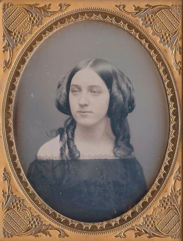 A vintage portrait of a young woman with dark hair styled in ringlets, wearing an off-the-shoulder dark dress, framed in an ornate gold oval frame decorated with floral patterns.