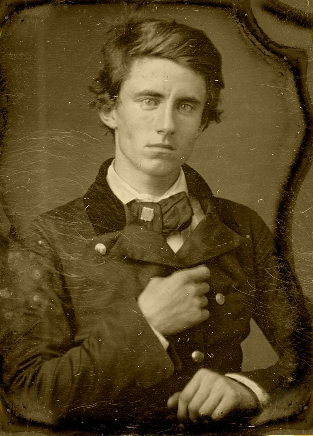 Sepia-toned portrait of a young man with light eyes and tousled hair, wearing a dark double-breasted coat and bow tie, gazing seriously at the camera with one hand partially tucked into his coat.