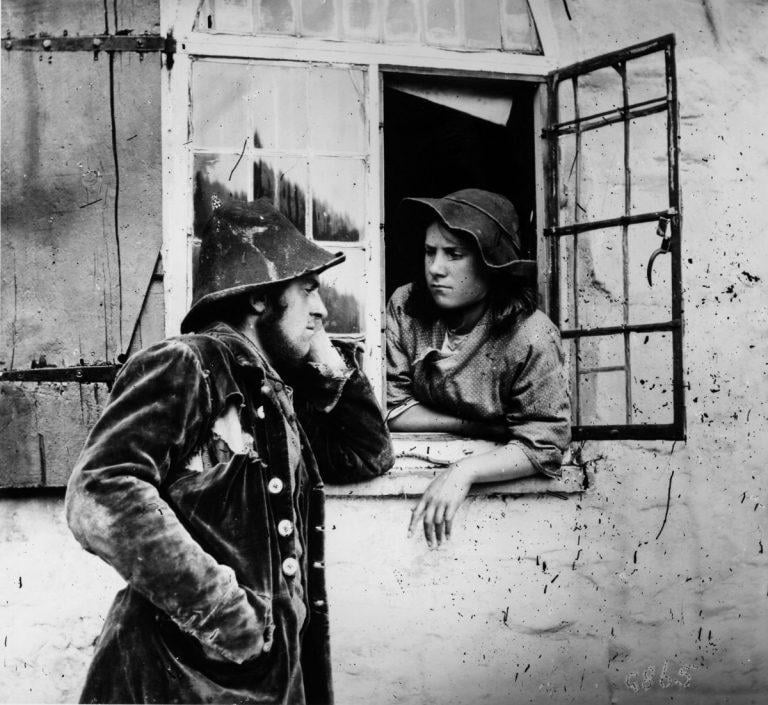 A black-and-white photo of a man in a coat and hat leaning on a windowsill, speaking with a woman in a bonnet who is leaning out of an open window, both wearing old-fashioned clothing.