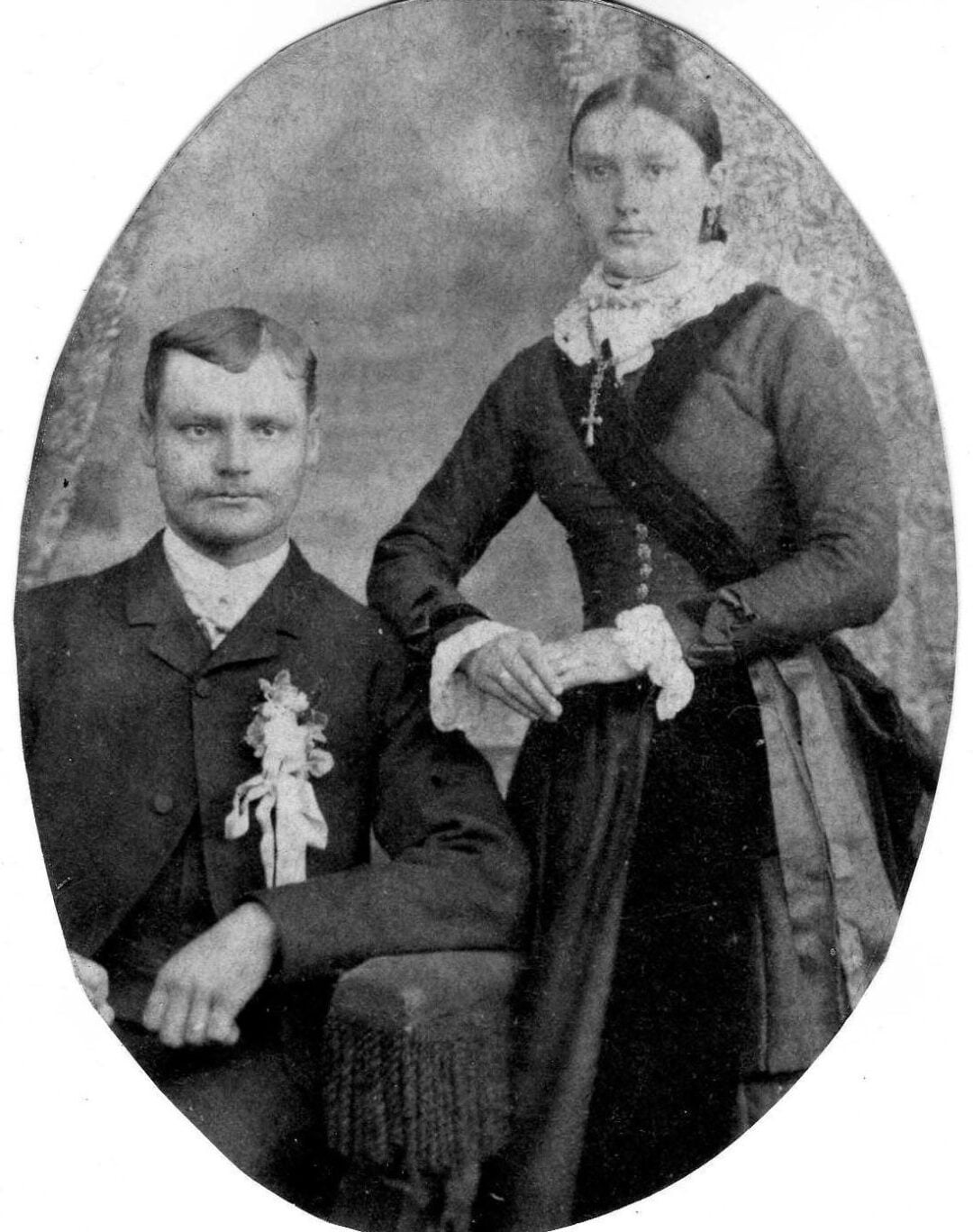 Black and white vintage portrait of a seated man in a suit with a boutonnière and a standing woman in a long dress, lace collar, and cross necklace, holding a folded item, likely from the late 19th or early 20th century.