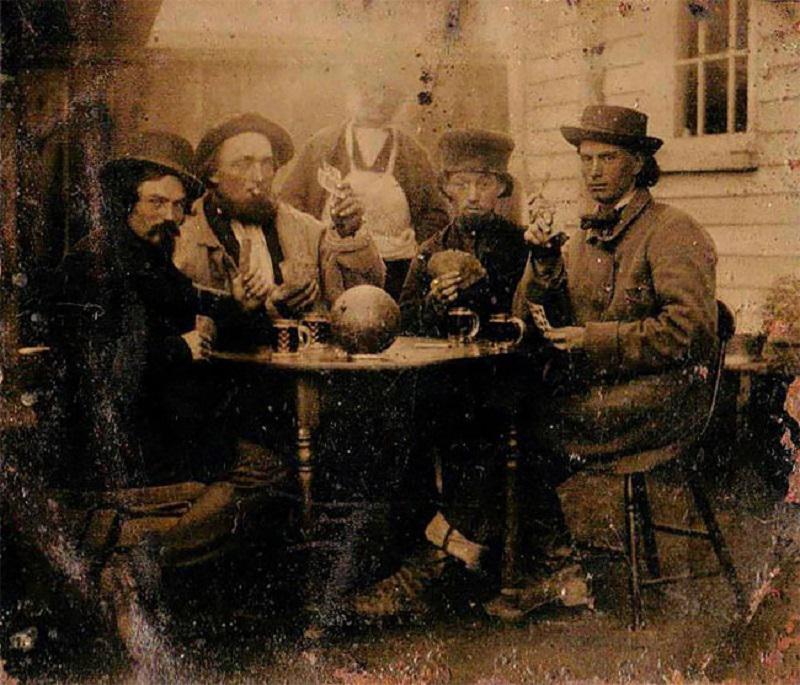 Five men in 19th-century attire sit around a table outside, smoking pipes and holding mugs. An additional person stands behind them. The scene appears sepia-toned and aged, suggesting an old photograph.