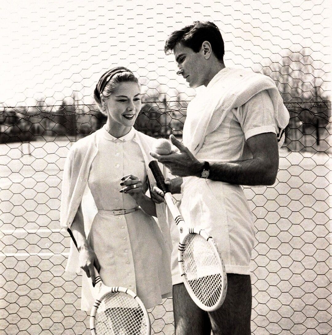 A woman and a man in vintage tennis outfits stand on a tennis court, holding racquets and a tennis ball, smiling and conversing near the net. The image is in black and white.