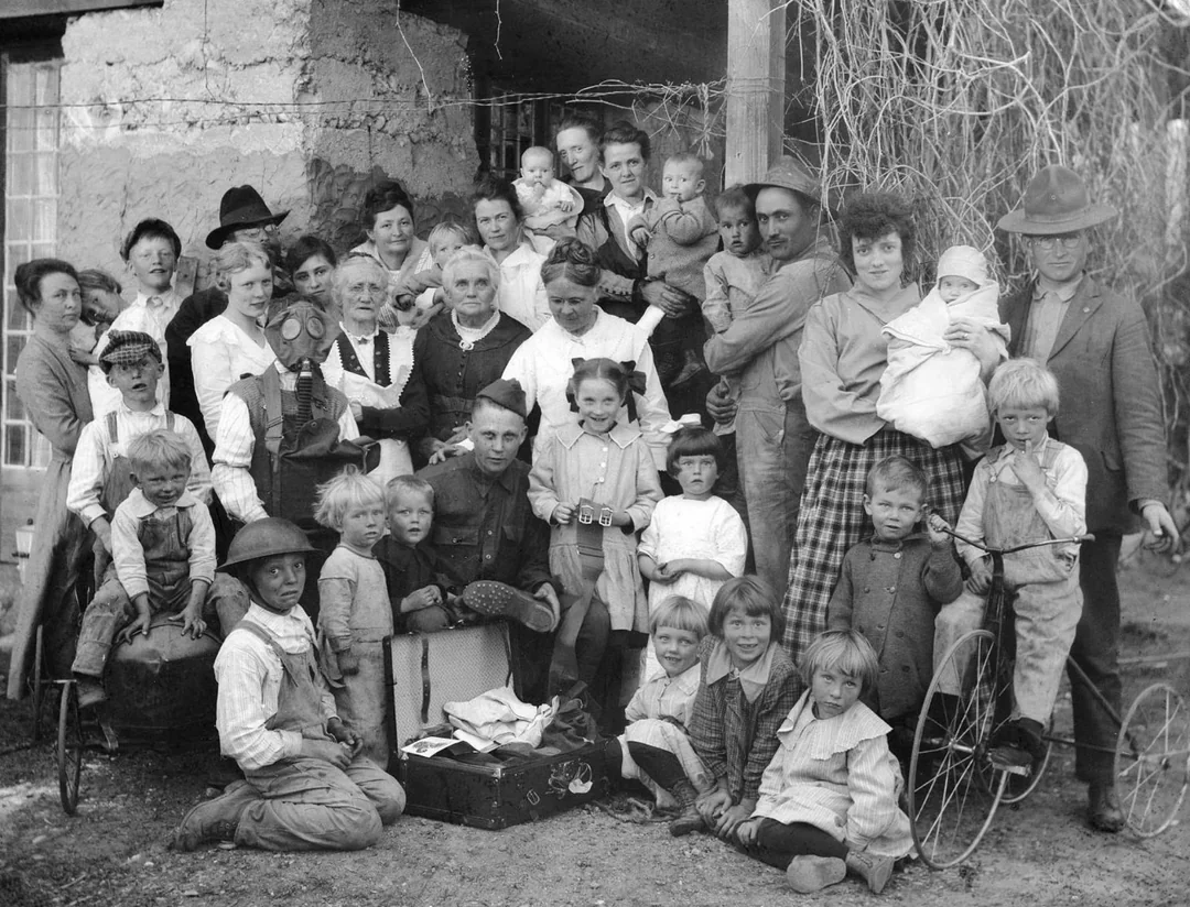 A large group of men, women, and children pose outside a rustic house. Some adults hold babies, and several children gather around a box with items. Old-fashioned clothing and a tricycle suggest the photo is from the early 1900s.