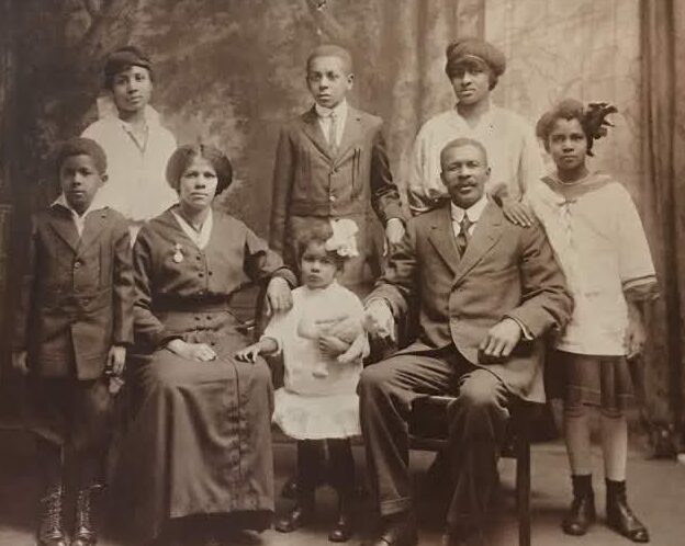 A vintage black-and-white photo of a family with four children standing and a man and woman seated, posing with a small child, all dressed in early 20th-century clothing, against a studio backdrop.