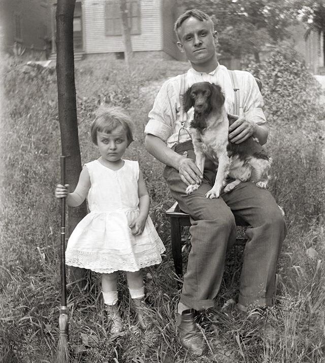 A man sits on a chair outdoors holding a dog, while a young girl in a white dress stands beside him holding a long rifle. They are in a grassy yard with houses and trees in the background.