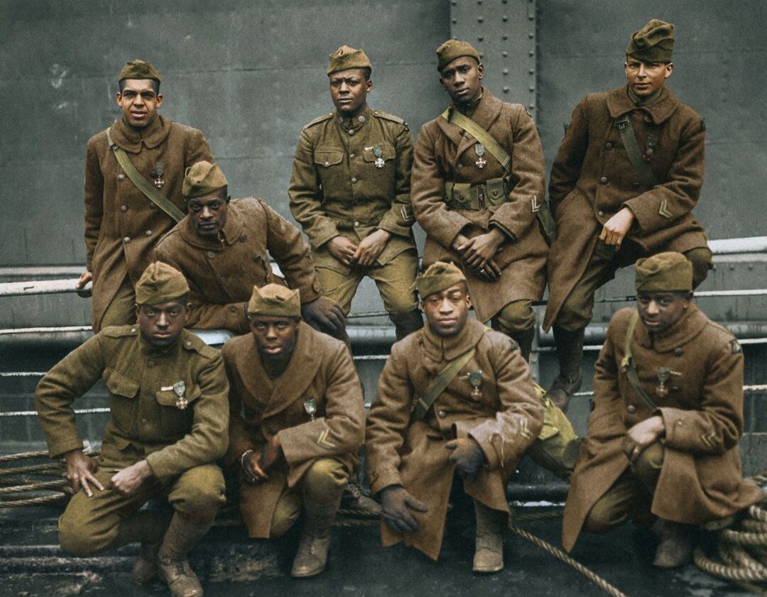 Nine African American soldiers in World War I uniforms pose together, some sitting and some standing, against an industrial backdrop. Most display medals on their coats and wear brown military attire and caps.