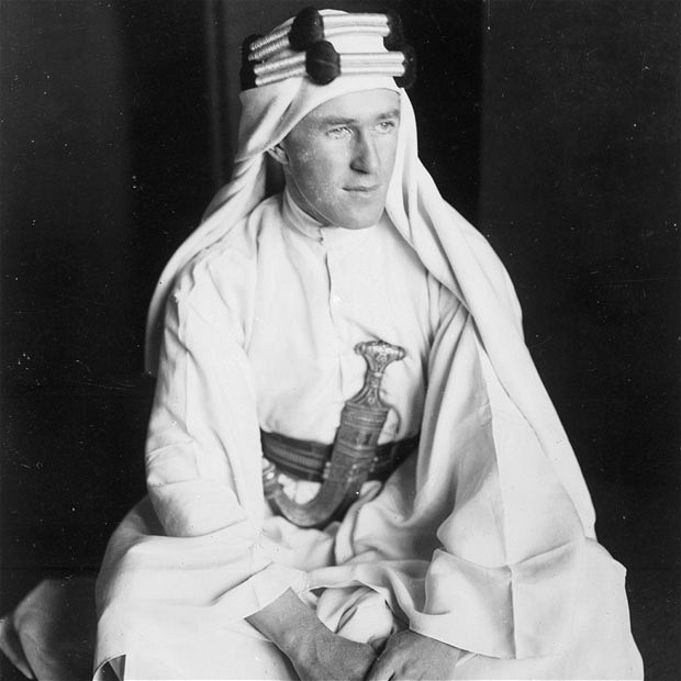 A man in traditional Middle Eastern attire, including a robe, belt, and a headscarf with cords, sits and looks to the side. A decorative curved dagger is tucked into his belt. The photo is in black and white.