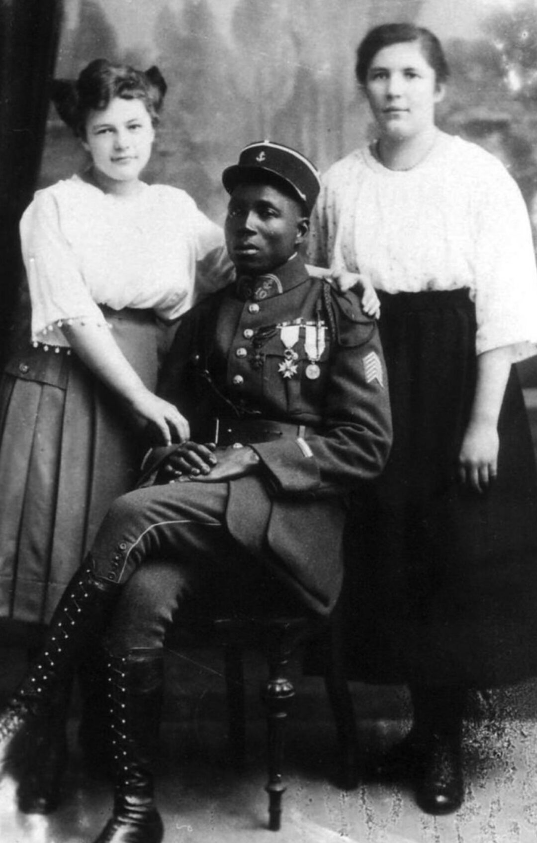 Black-and-white photo of a decorated soldier in uniform sitting on a chair, flanked by two women in blouses and skirts, both resting their hands on his shoulders. They pose in front of a painted studio backdrop.