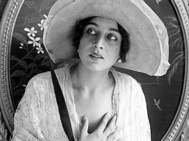 A woman in vintage clothing and a wide-brimmed hat looks upward with a thoughtful expression, one hand placed on her chest, in front of a decorative, floral-patterned background.