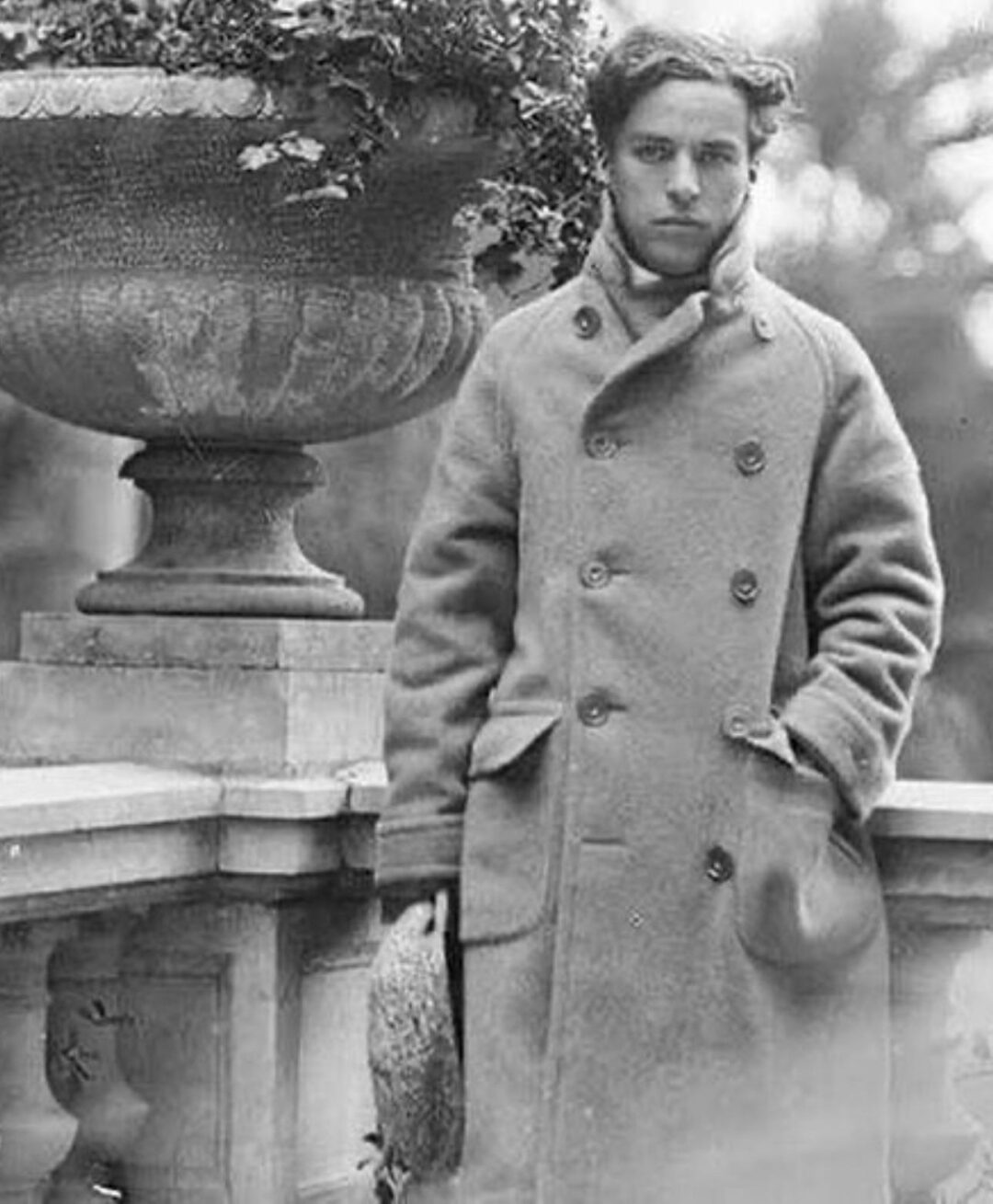 A young man with wavy hair wearing a long, double-breasted overcoat stands by a stone railing and large planter, looking at the camera with one hand in his pocket. The image is in black and white.