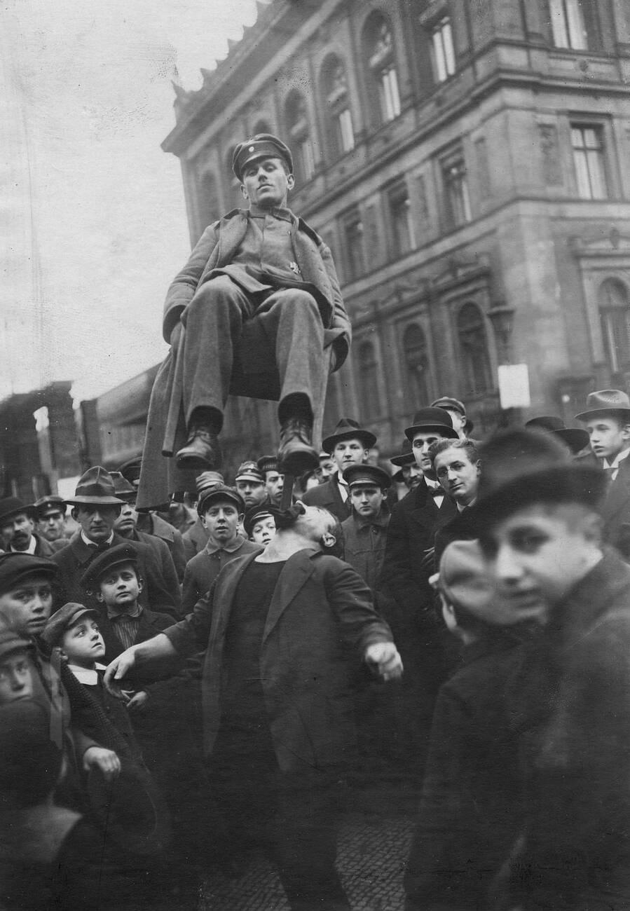 A man balances another man sitting on a chair on his chin, surrounded by a crowd of onlookers in old-fashioned clothing, with a large stone building in the background.