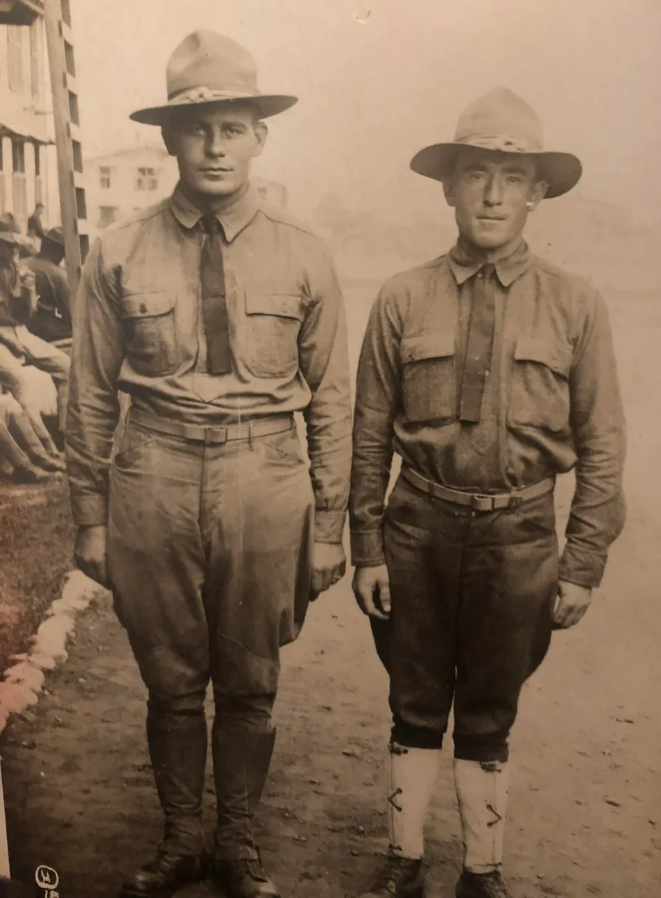 Two men in early 20th-century military uniforms and wide-brimmed hats stand side by side outdoors, with buildings and several people seated in the background. The photo is black and white.