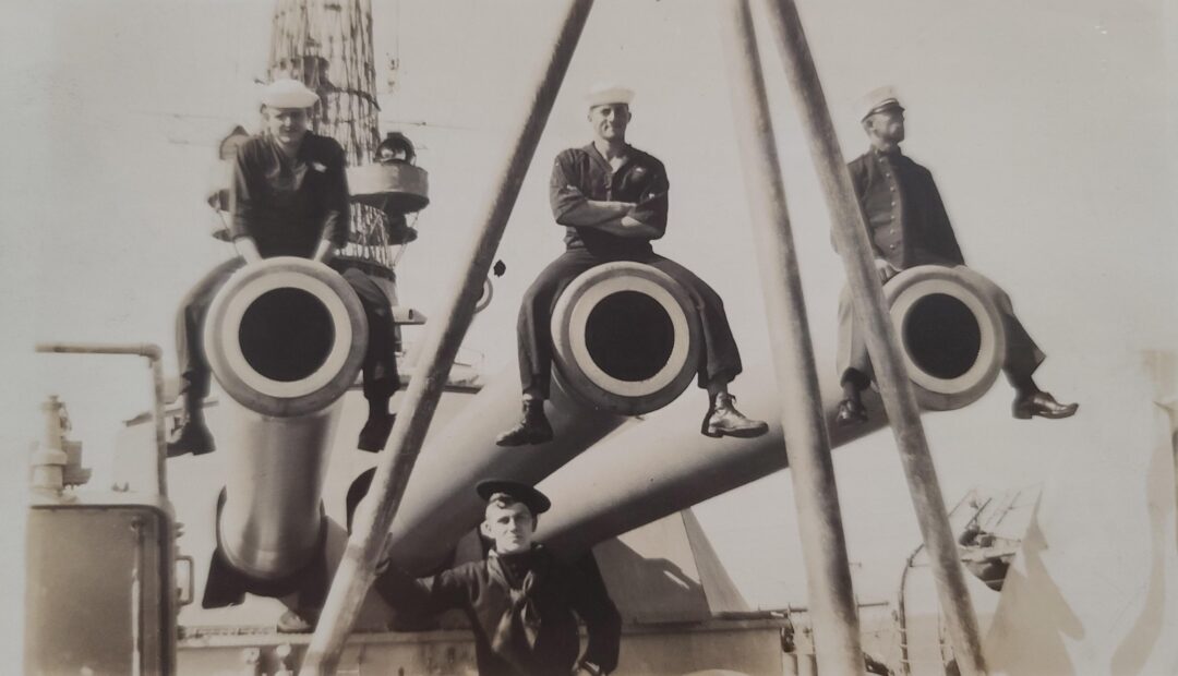 Four sailors pose on a navy ship; three sit atop large gun barrels while one stands below, all wearing uniforms and hats, with ship equipment visible in the background.