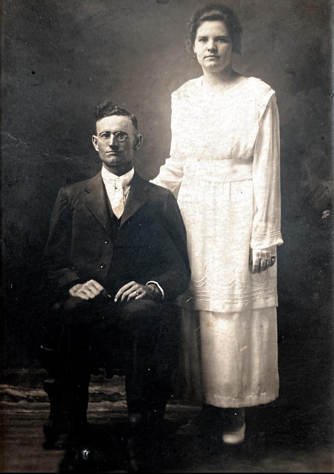 A vintage black-and-white photo of a man seated in a suit and glasses, with a woman standing beside him, resting her hand on his shoulder. She wears a long, light-colored dress. Both look seriously at the camera.