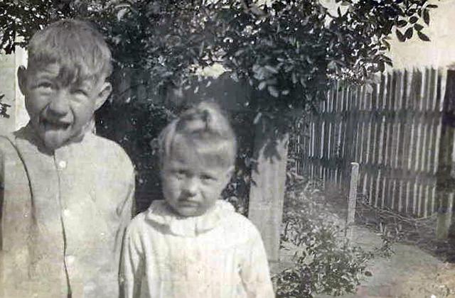 Two young children stand outdoors near a wooden fence and leafy tree. The child on the left makes a funny face, sticking out their tongue, while the other looks at the camera with a neutral expression. The photo is in black and white.