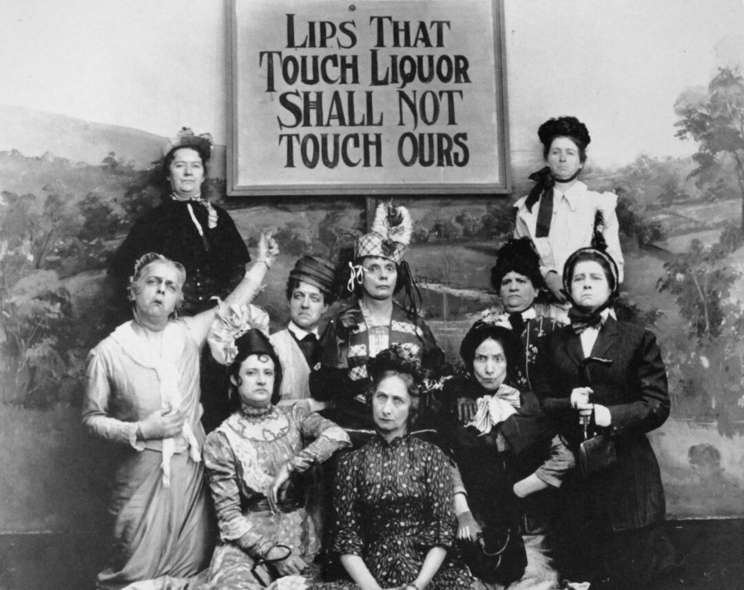 A group of stern-faced women in old-fashioned clothes pose beneath a sign reading, “Lips That Touch Liquor Shall Not Touch Ours.” The background is a painted countryside scene.
