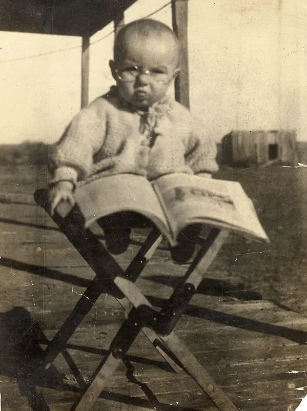 A sepia-toned vintage photo of a baby sitting on a folding chair outdoors, wearing a knitted sweater and looking at the camera with a serious expression, with an open book on their lap.
