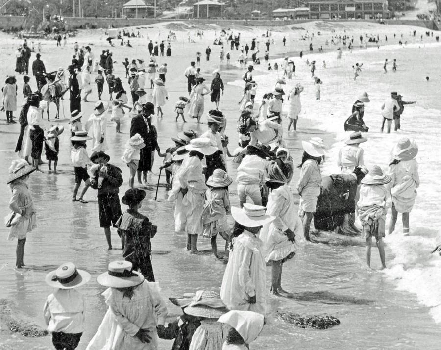 Crowds of people, mostly women and children in old-fashioned clothing and hats, stand and wade in the shallow water at a busy beach on a sunny day, with more people visible along the shoreline in the background.