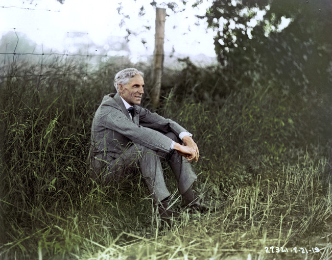 An older man in a grey suit sits on the grass by a wire fence, surrounded by tall grass and greenery, smiling and looking to his left. The photo appears vintage and colorized.