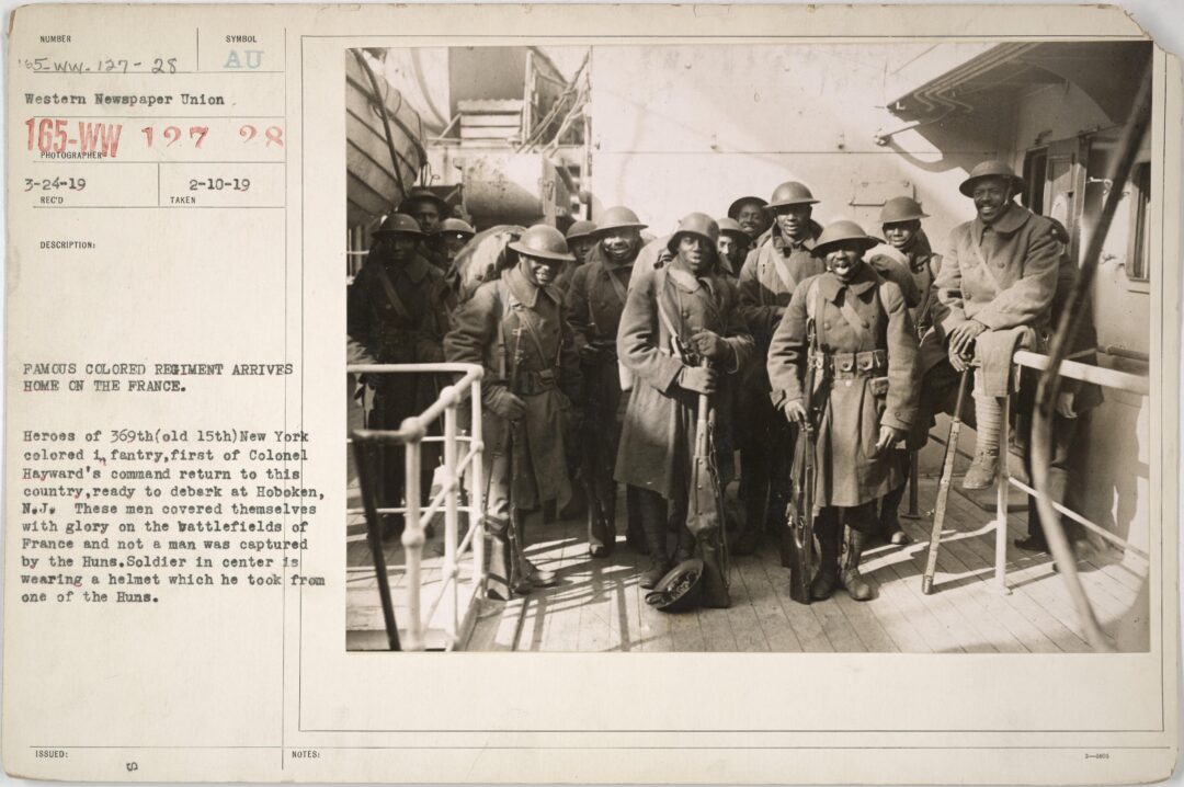 A group of African American soldiers in uniform and helmets stand smiling on a ship's deck during World War I, with text identifying their arrival home after being captured by German forces.